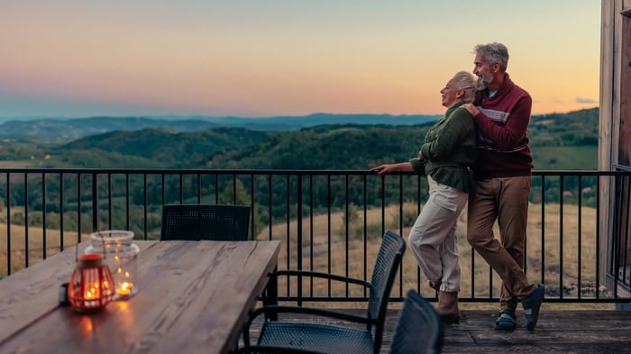 Dos personas apoyadas en la barandilla de una terraza de madera, mirando un paisaje de colinas al atardecer, con una mesa y velas encendidas en primer plano