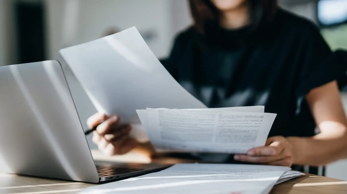 Persona revisando documentos impresos junto a un ordenador portátil en una mesa de trabajo