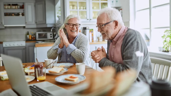 Dos personas adultas sentadas en una mesa de cocina con un ordenador portátil y desayuno, en un entorno doméstico luminoso