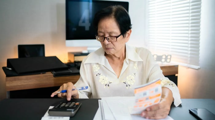 Persona mayor sentada en una mesa revisando documentos y usando una calculadora en un espacio de trabajo en casa