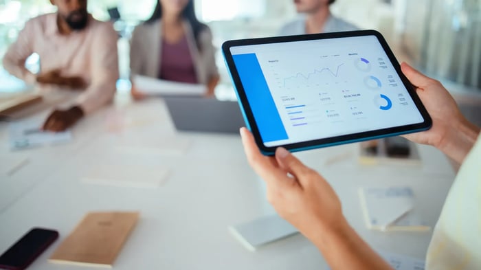 reunión de personas en una sala, con fondo difuminado y unas manos sujetando una tablet con un gráfico