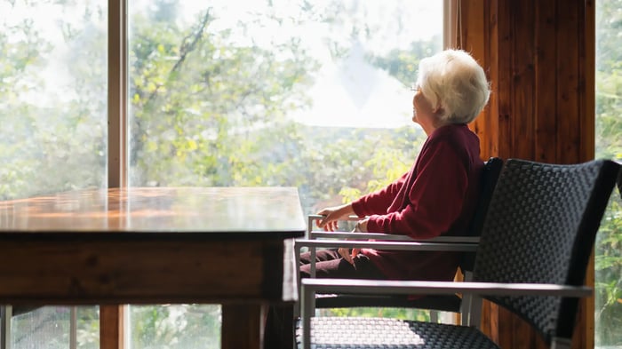 Persona mayor sentada en una silla junto a una mesa, mirando hacia una ventana con vistas a un entorno natural